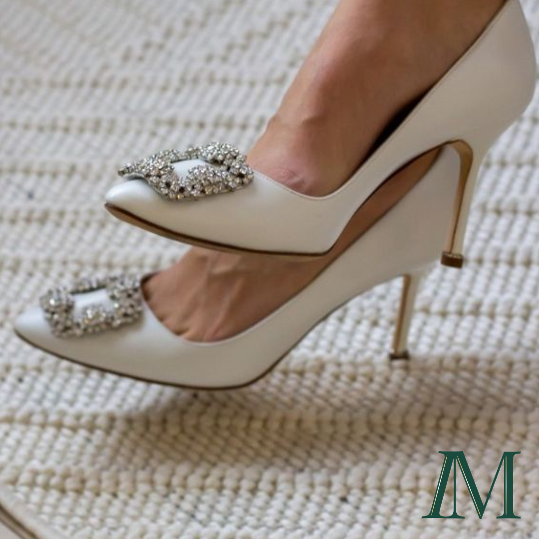 The image of a pair of women's feet, crossed at the ankle, wearing a beige pair of Manolo Blahnik Hangisi crystal buckle high heel shoes. There is beige carpet on the floor, and the Inspired Method IM logo in green is in the lower right corner.